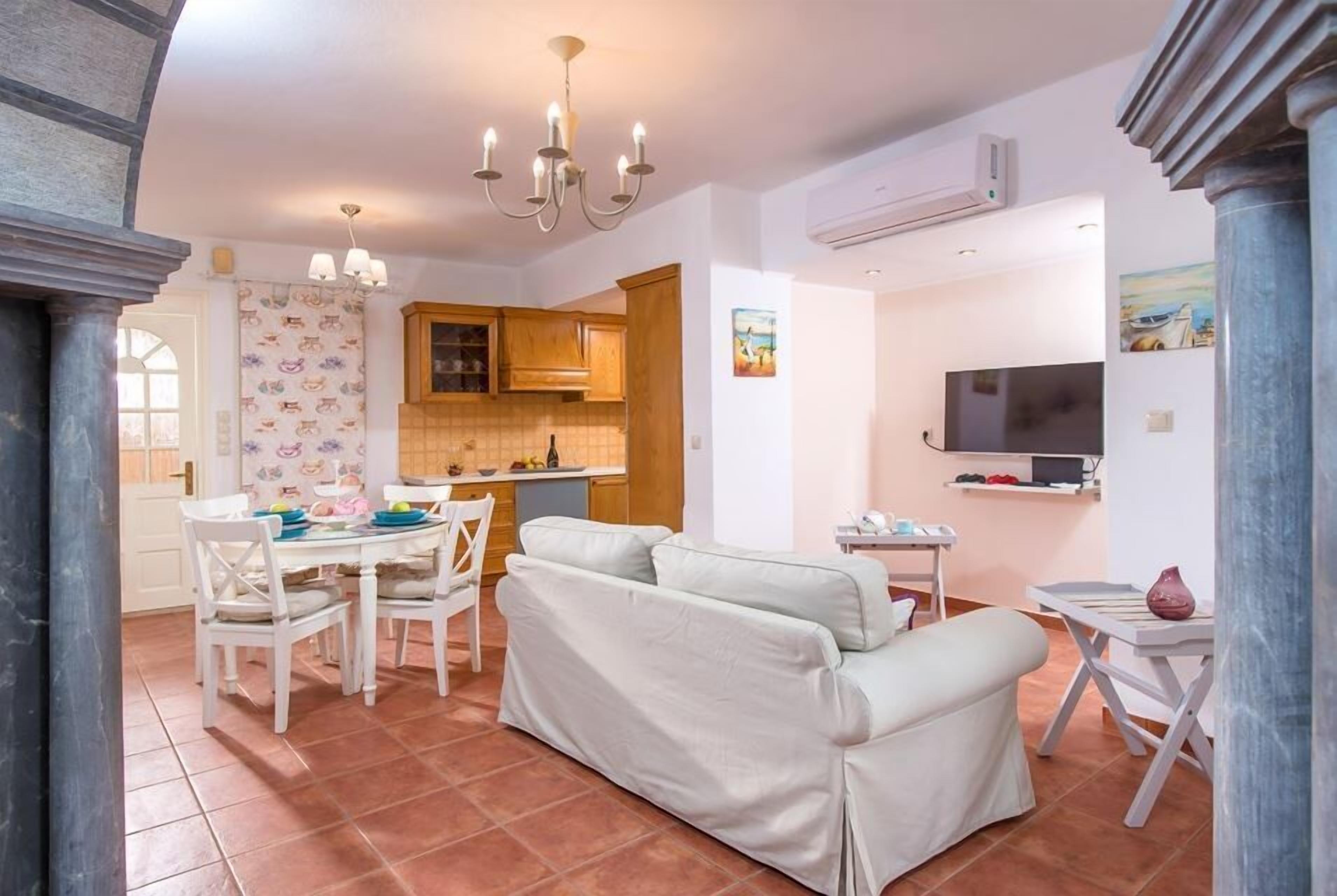 Una cuina moderna amb armaris blancs, una zona de menjador amb taula i cadires blanques, i un saló amb un sofà blanc.