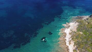 An aerial view of a coastal area with clear waters, rocky shoreline, and sparse vegetation.