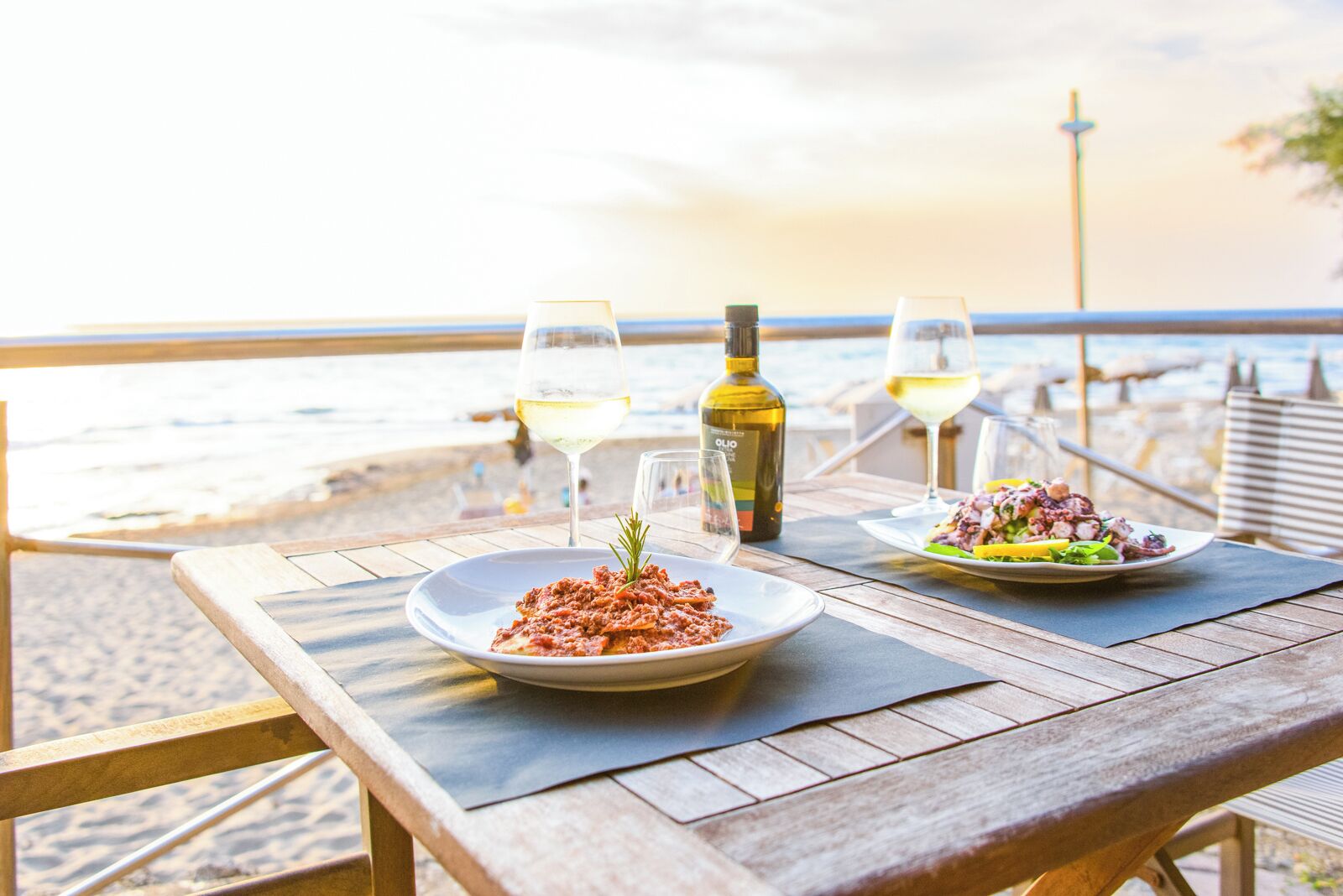 A wooden table with two plates of food and two glasses of wine on a beach overlooking the sea.