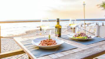 A wooden table with two plates of food and two glasses of wine on a beach overlooking the sea.