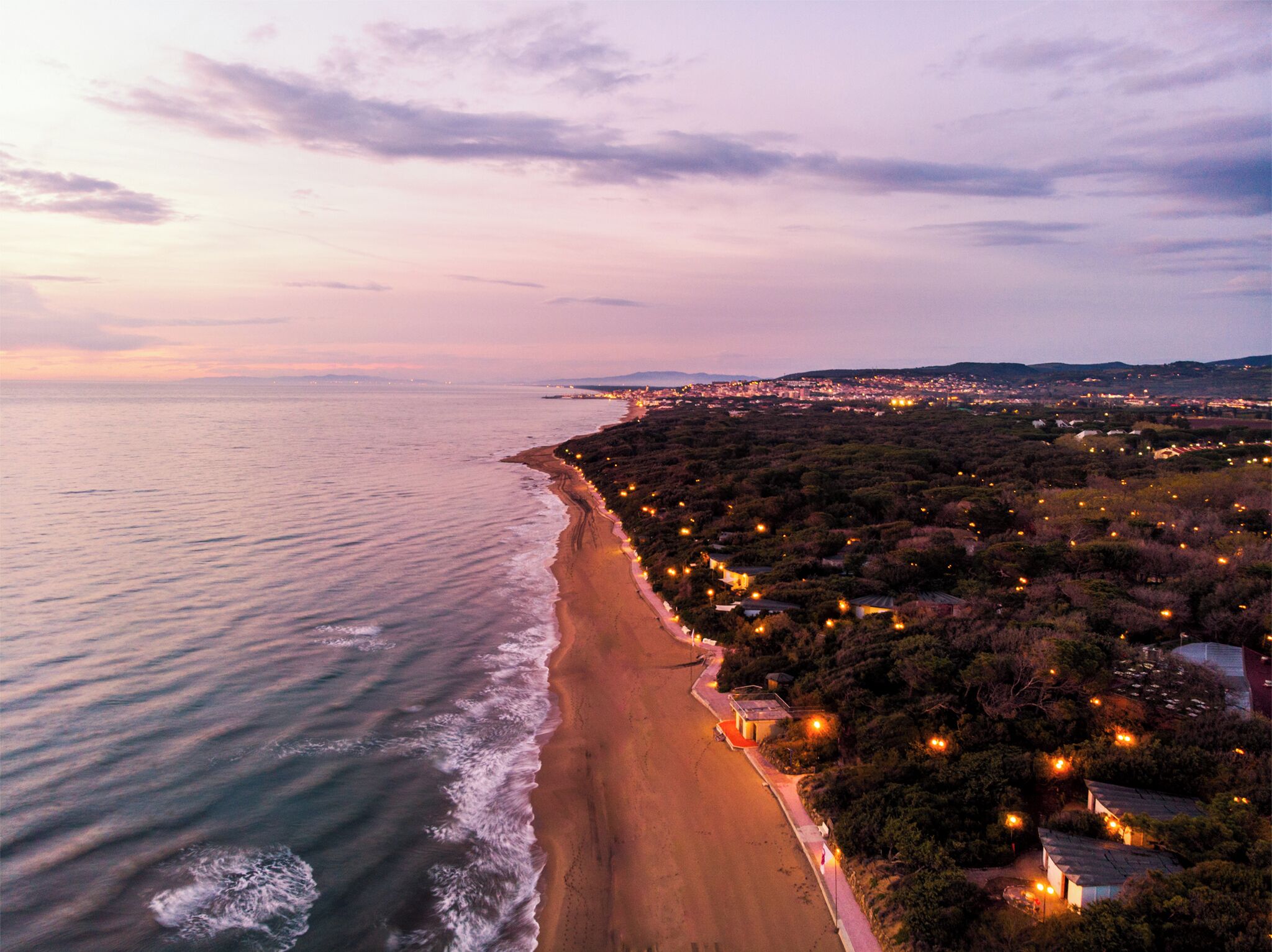 A coastal road with a beach on one side and a densely wooded area on the other, with streetlights illuminating the path.