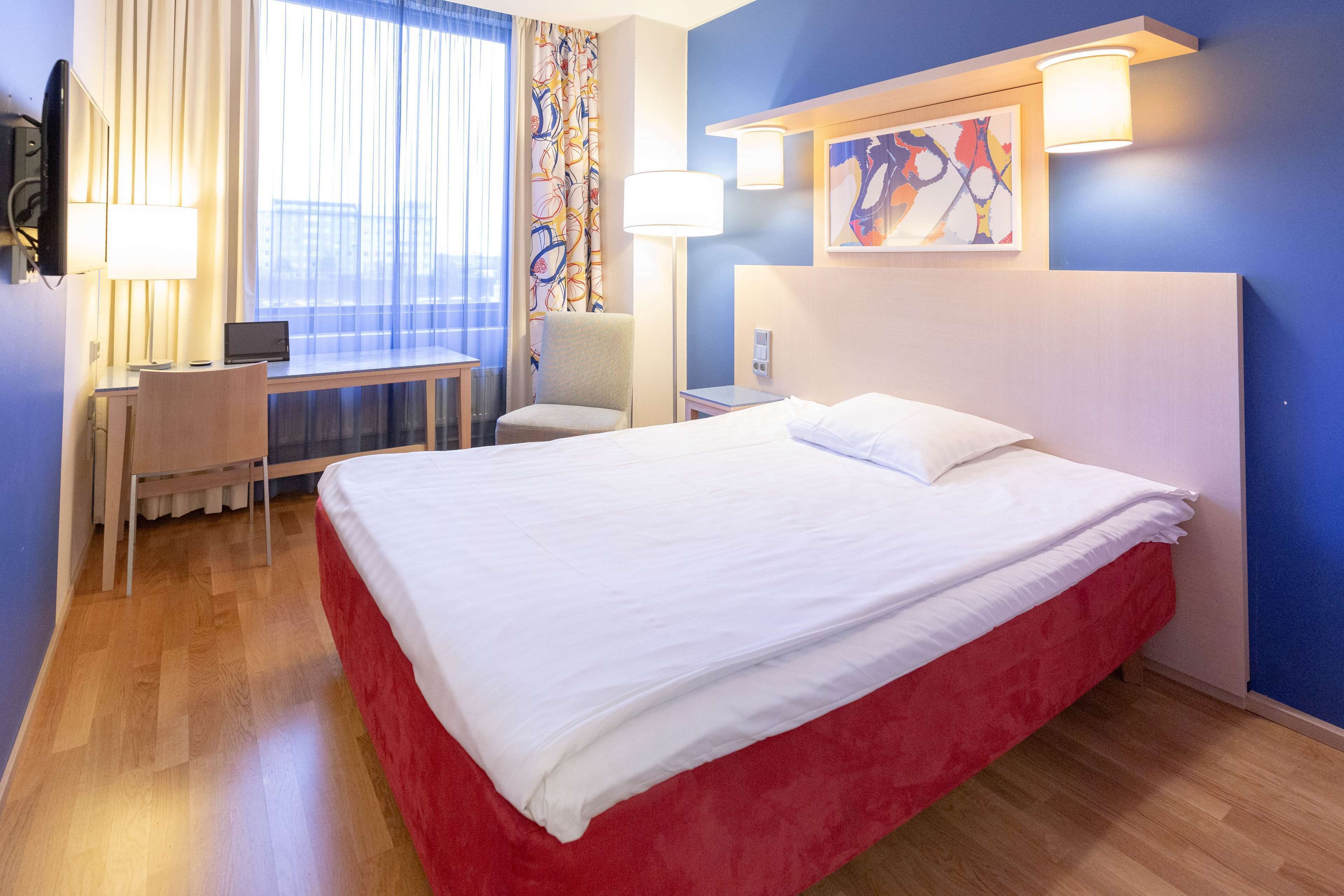 A hotel room with a large bed, a desk with a laptop, a chair, and a window with curtains.