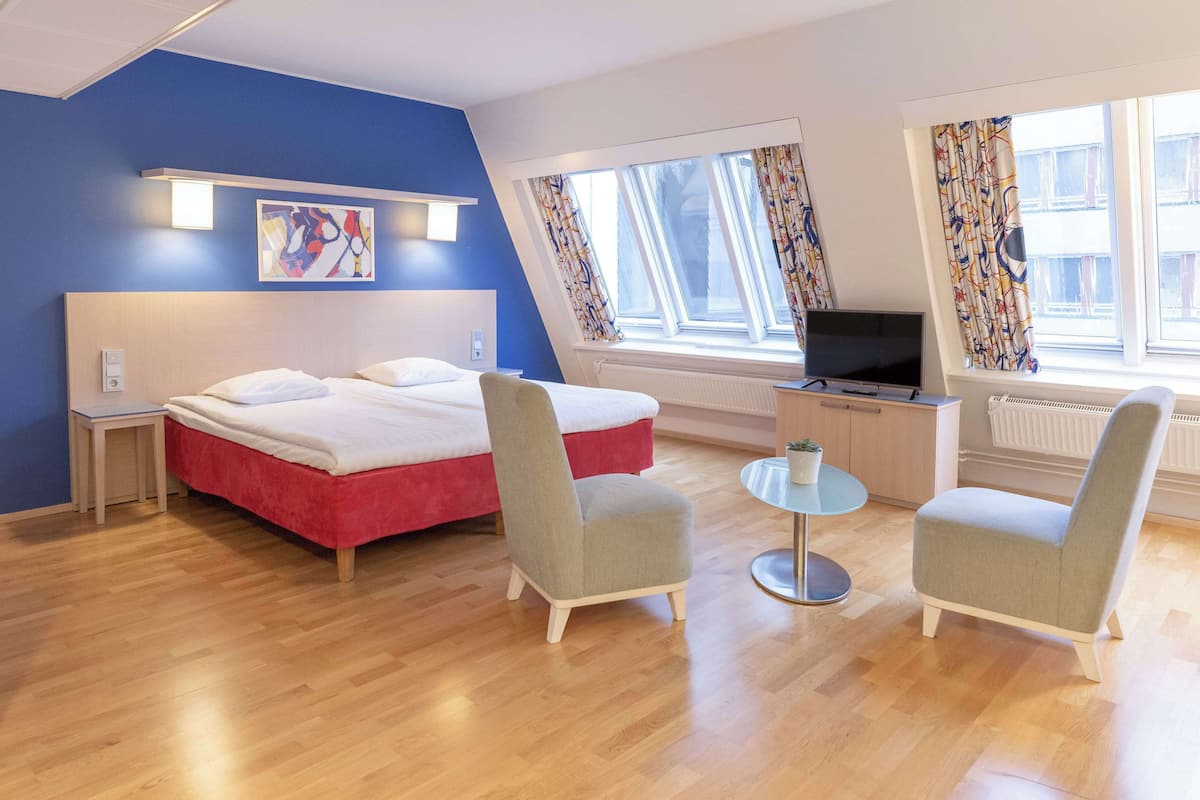 A modern hotel room with a bed, two armchairs, a TV, and large windows with curtains.