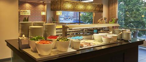 A buffet setup with various food items and utensils, a patterned wall, and a window with a view of trees.