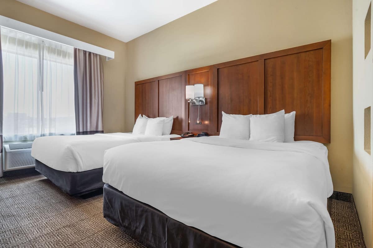 Habitación de hotel con dos camas, un cabecero de madera grande, una mesita de noche con lámpara y un ventanal con cortinas.
