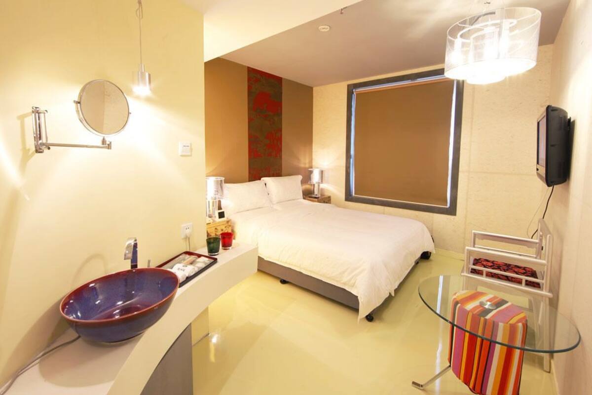 A modern hotel room with a large bed, a round mirror, a bowl-shaped sink, and a glass table with a striped bag.