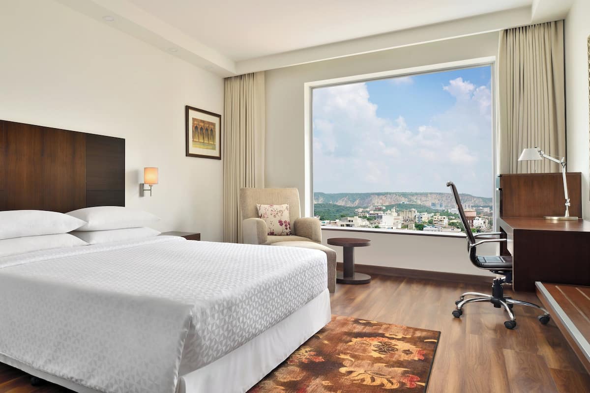 Una habitación de hotel con una cama grande, un escritorio, una silla, una mesita y vistas a la ciudad a través de un ventanal amplio.