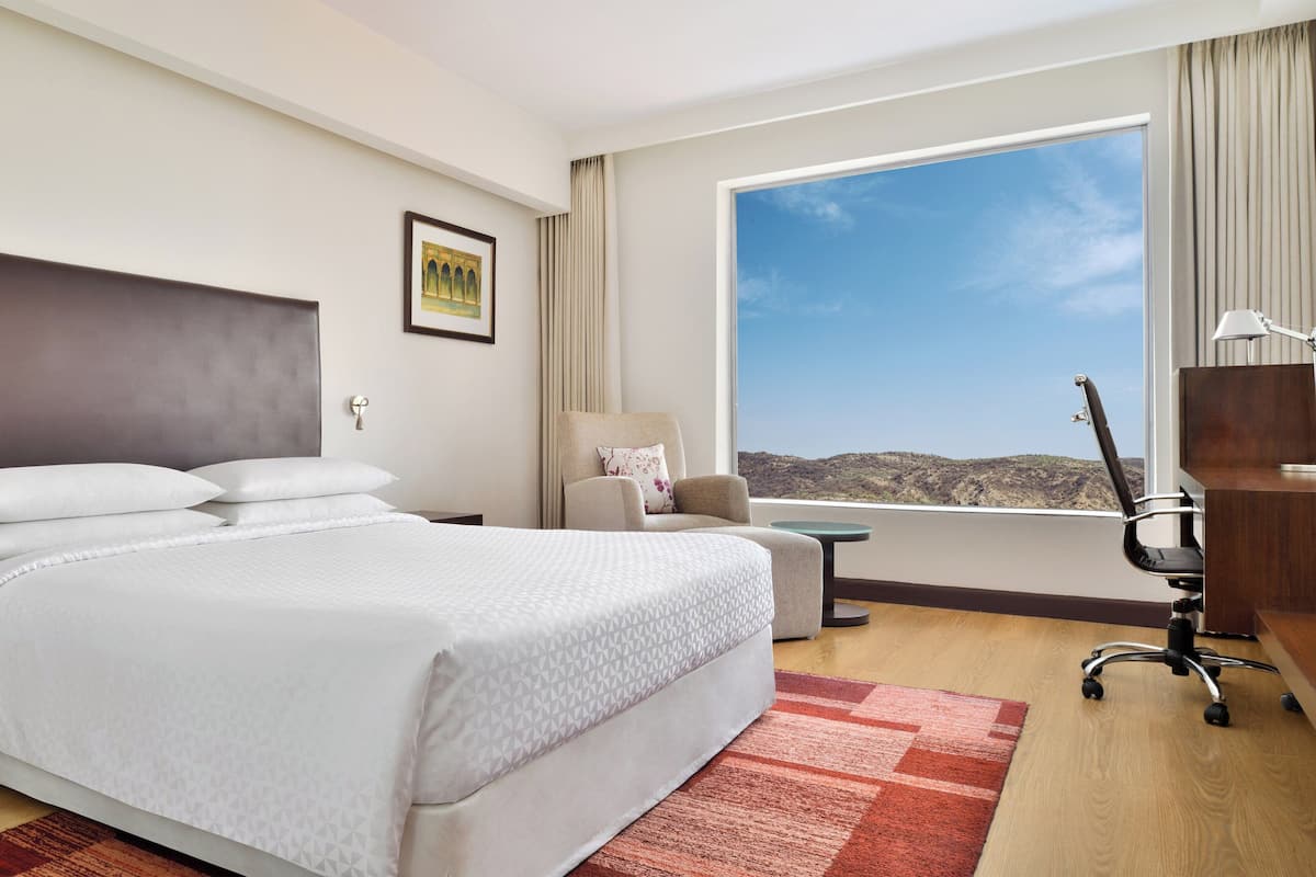 Habitación de hotel con una cama grande, un escritorio, una silla y vistas a las montañas a través de un ventanal amplio.