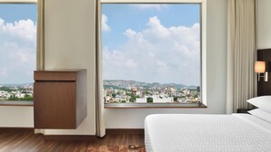 Une chambre d’hôtel dotée d’une grande fenêtre donnant sur la ville, d’un lit, d’une table de chevet et d’un plancher en bois.