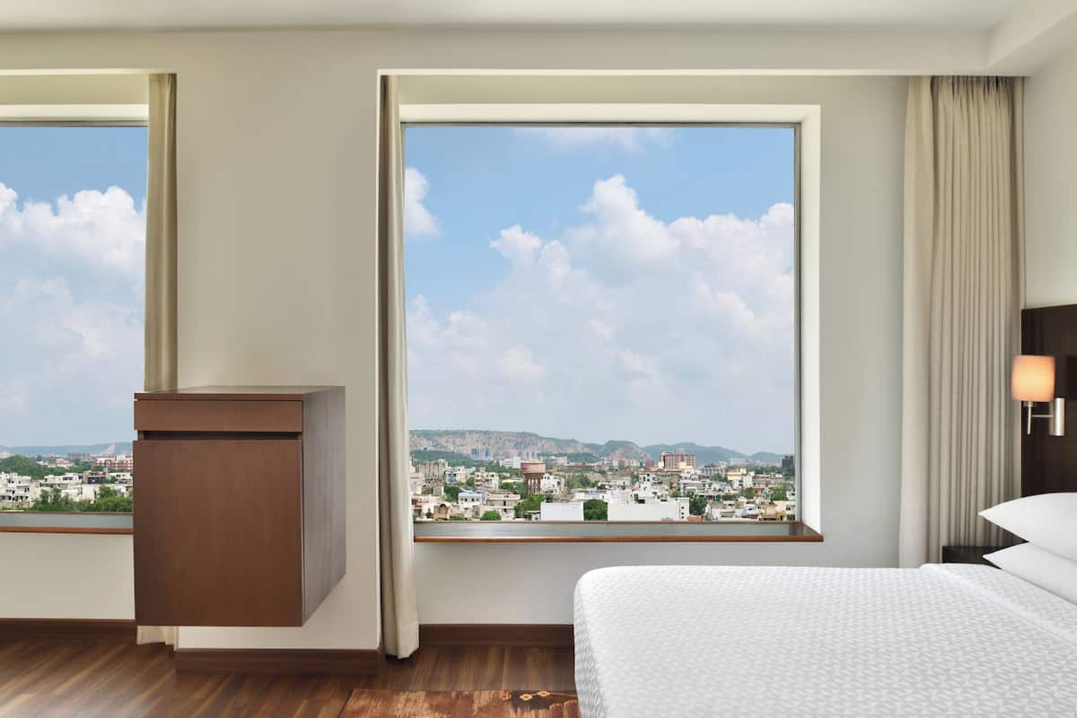 Habitación de hotel con un ventanal grande que ofrece vistas a la ciudad, una cama, una mesita de noche y suelo de madera.