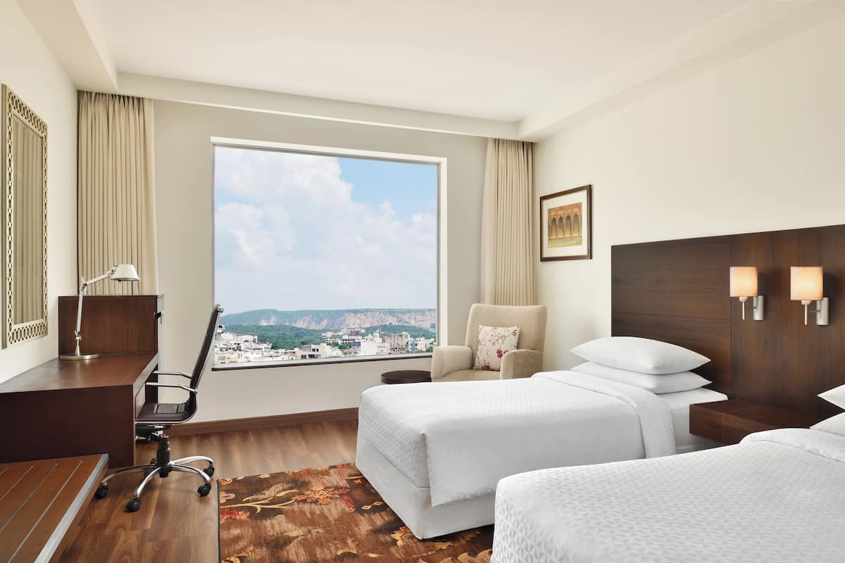 Habitación de hotel con un ventanal grande, escritorio, dos camas, una silla y mesita de noche con lámparas.
