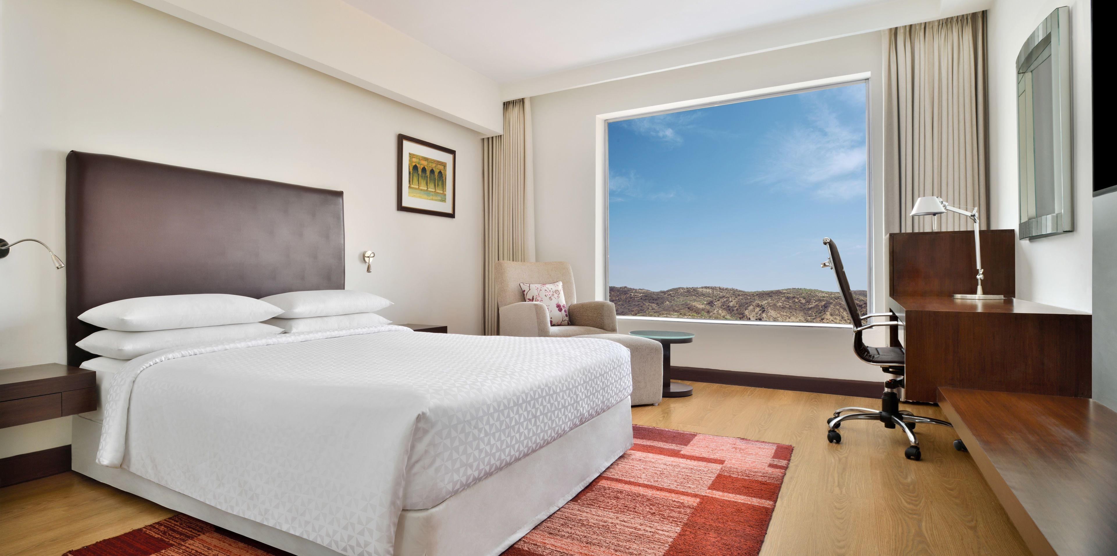 A hotel room with a large bed, a desk, a chair, a small table, and a window with a view of the hills.