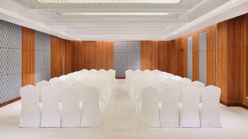 Une salle de conférence avec des rangées de chaises blanches, des murs lambrissés en bois et un sol à motifs.