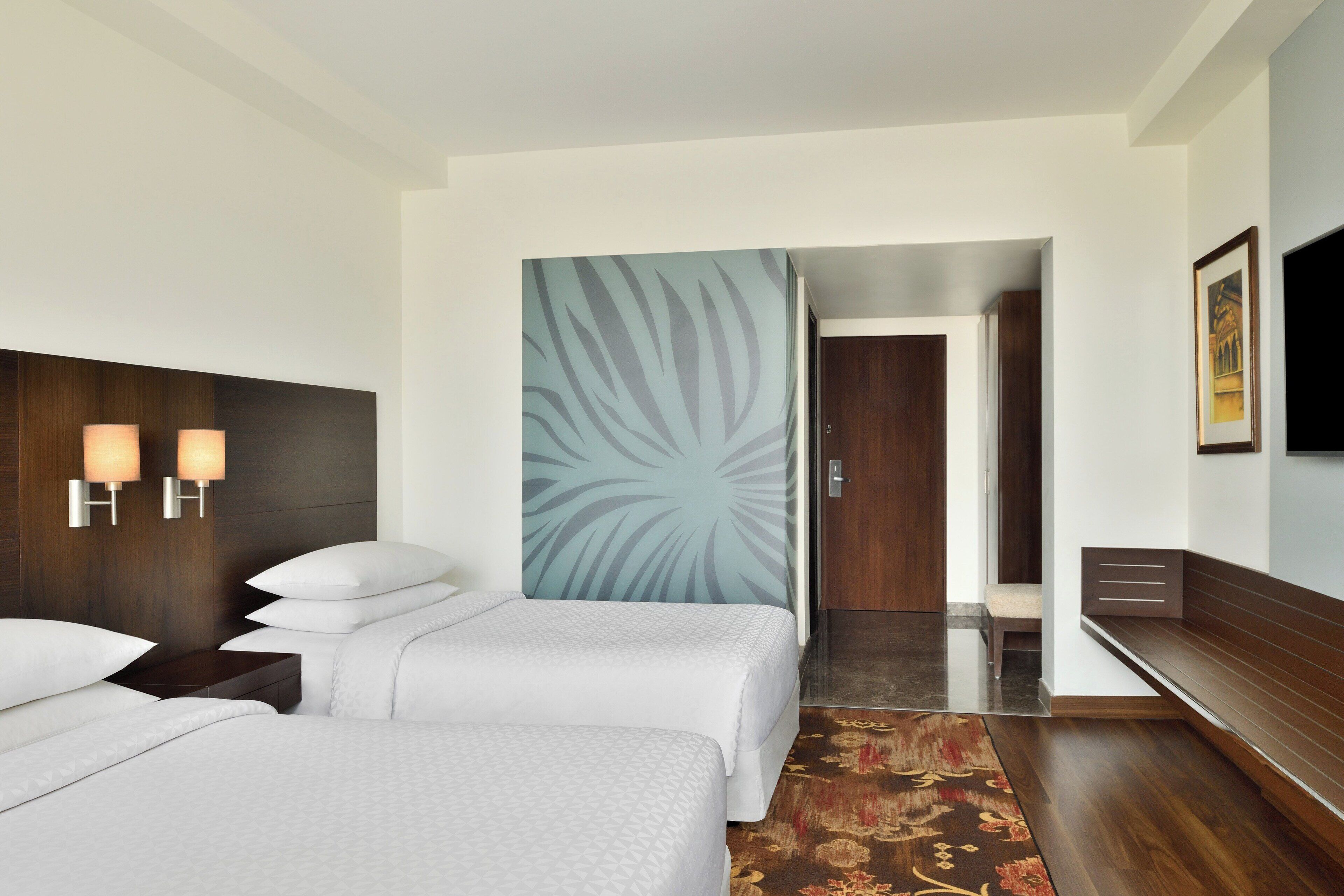 A hotel room with two beds, a large wall mural, a TV, and a wooden bench.