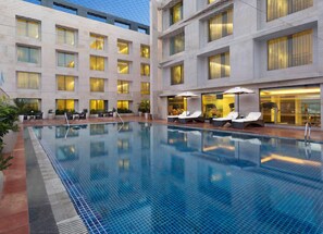 A hotel pool area with lounge chairs and umbrellas, surrounded by a building with multiple floors and windows.
