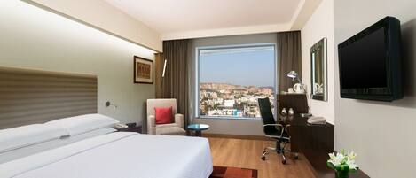 A hotel room with a large bed, a desk, a chair, a TV, and a view of a cityscape.