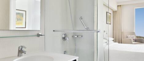 Un baño de hotel moderno con cabina de ducha de vidrio, un lavamanos blanco y una cama con vistas.