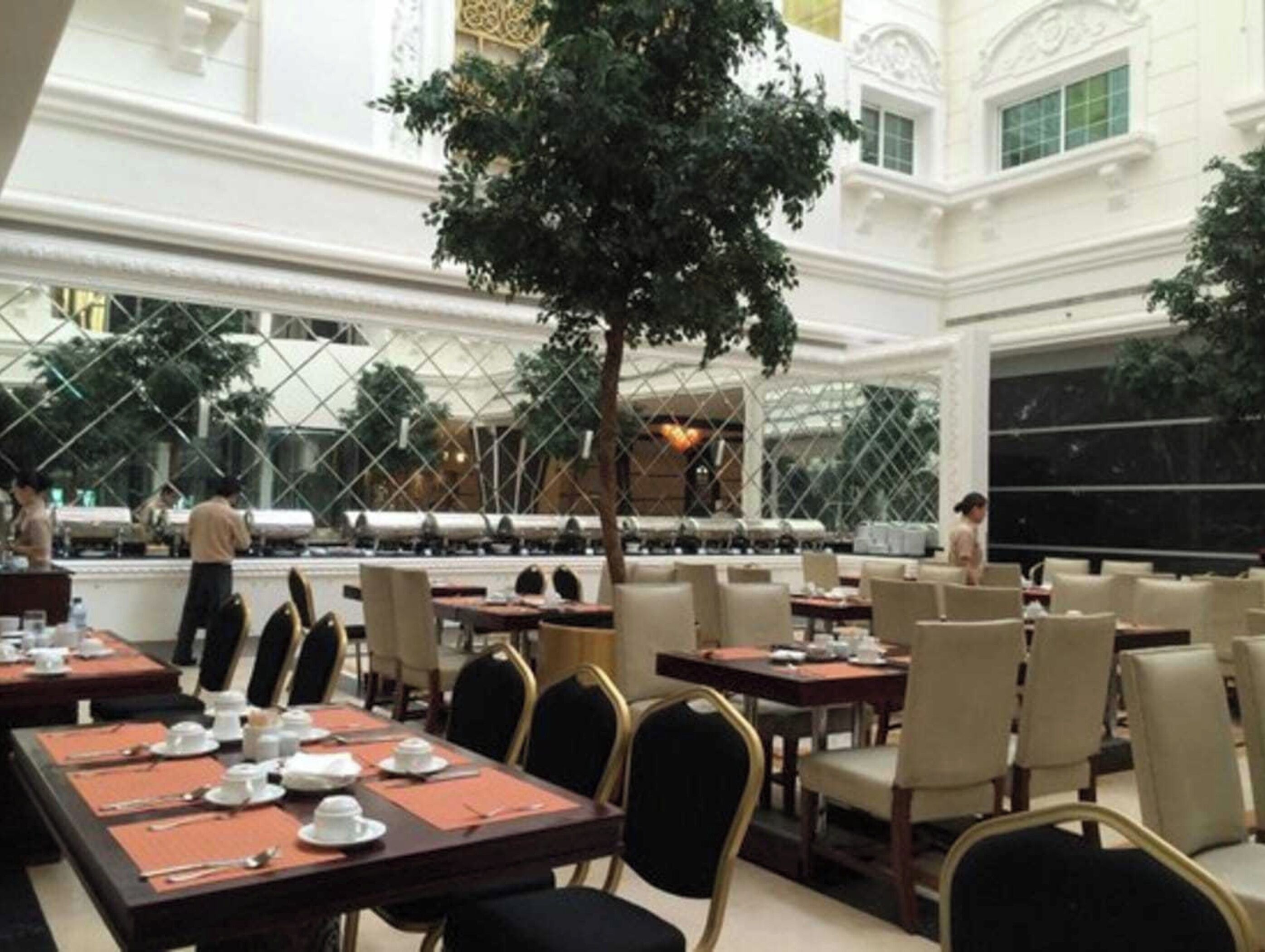 A spacious dining area with multiple tables and chairs, a large tree, and a view of the cityscape through large windows.