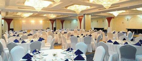 Un salón de banquetes con mesas redondas dispuestas para comer, sillas blancas y candelabros.