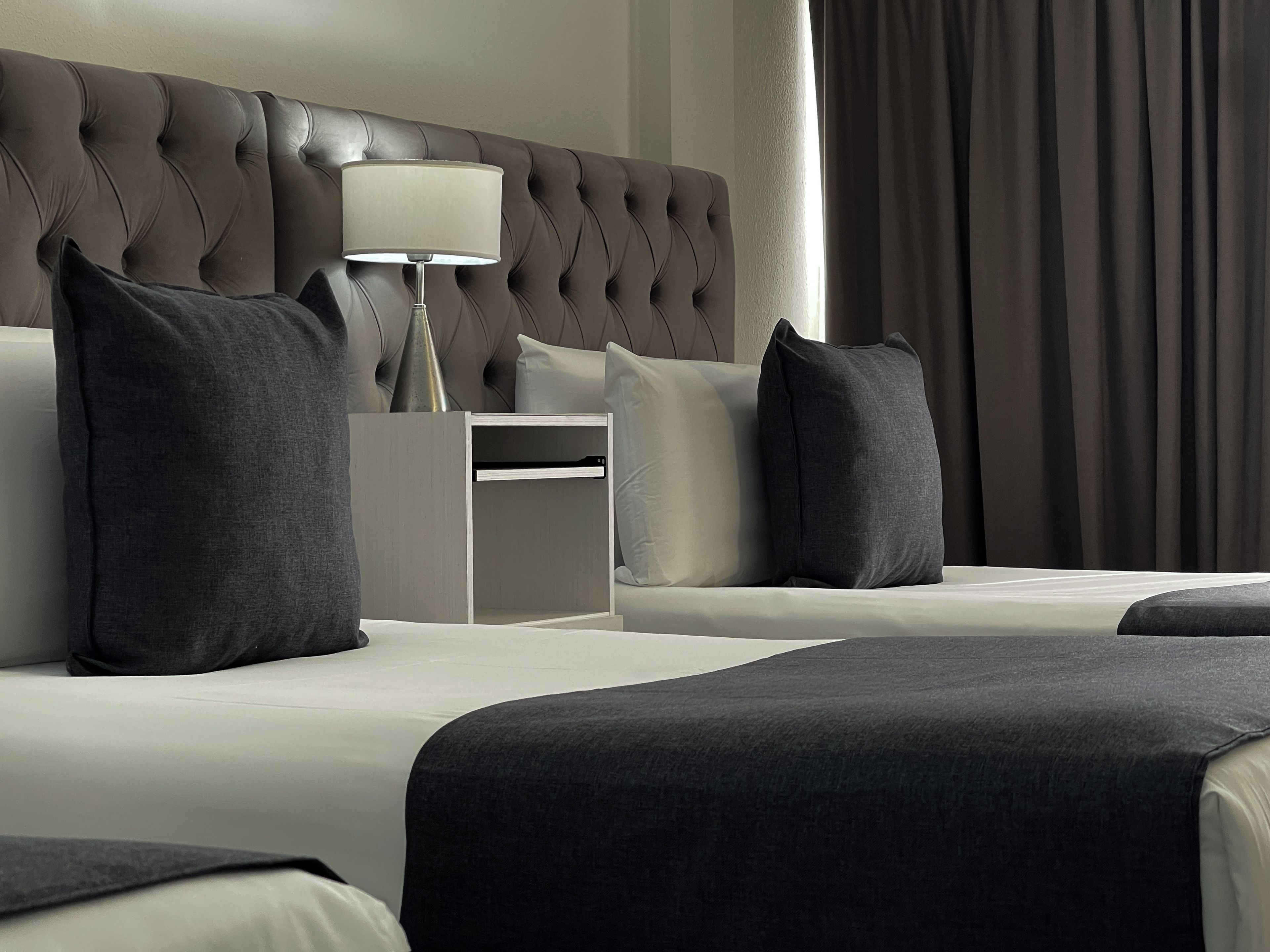 A hotel room with a tufted headboard, a bedside table with a lamp, and two beds with dark bedding.