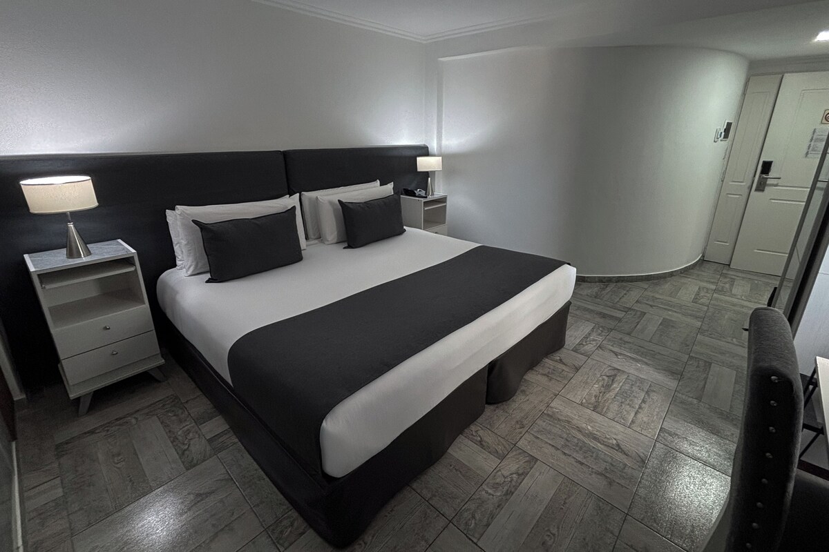 A modern hotel room with a large bed, two bedside tables, a nightstand, and a wardrobe.