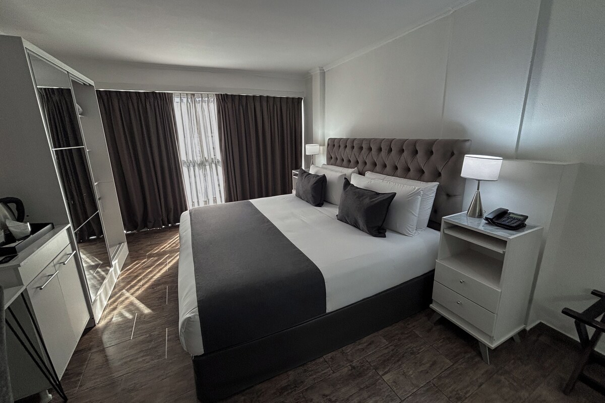 A modern hotel room with a large bed, a bedside table with a lamp, a telephone, and a wardrobe.