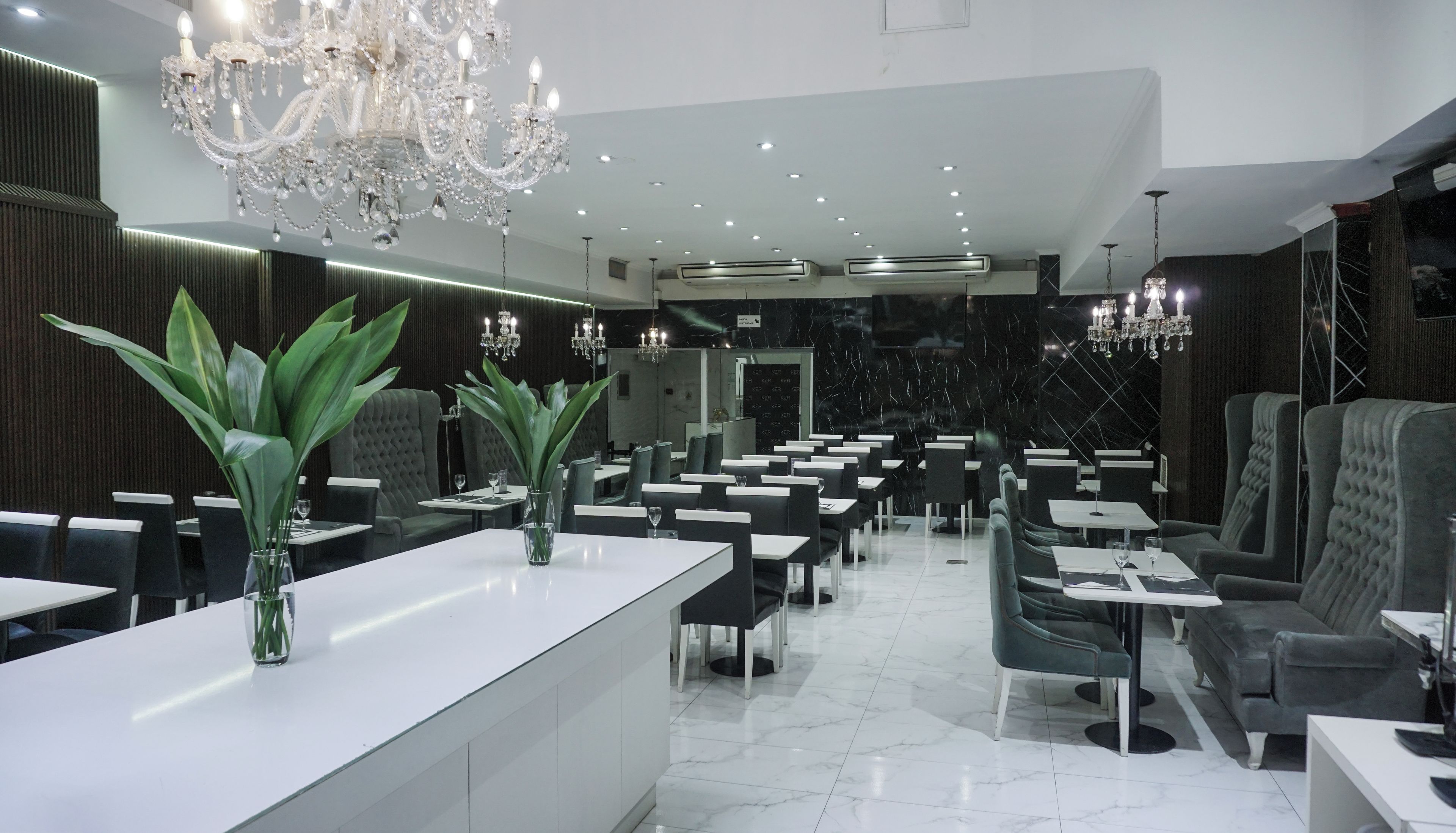 A modern dining area with a chandelier, marble flooring, and green plants.