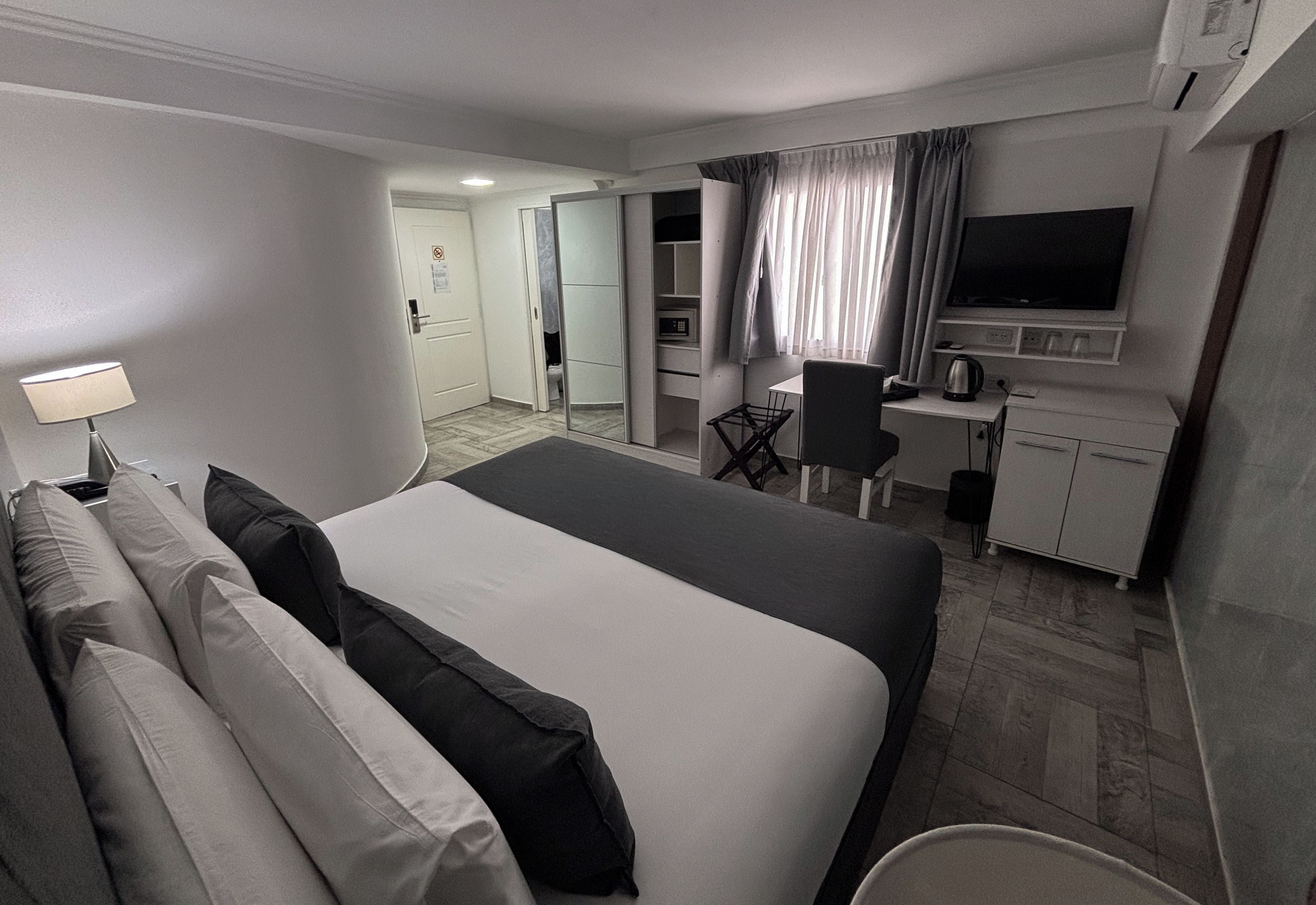 Quarto de hotel com cama, escrivaninha, cadeira, televisão e cozinha compacta.