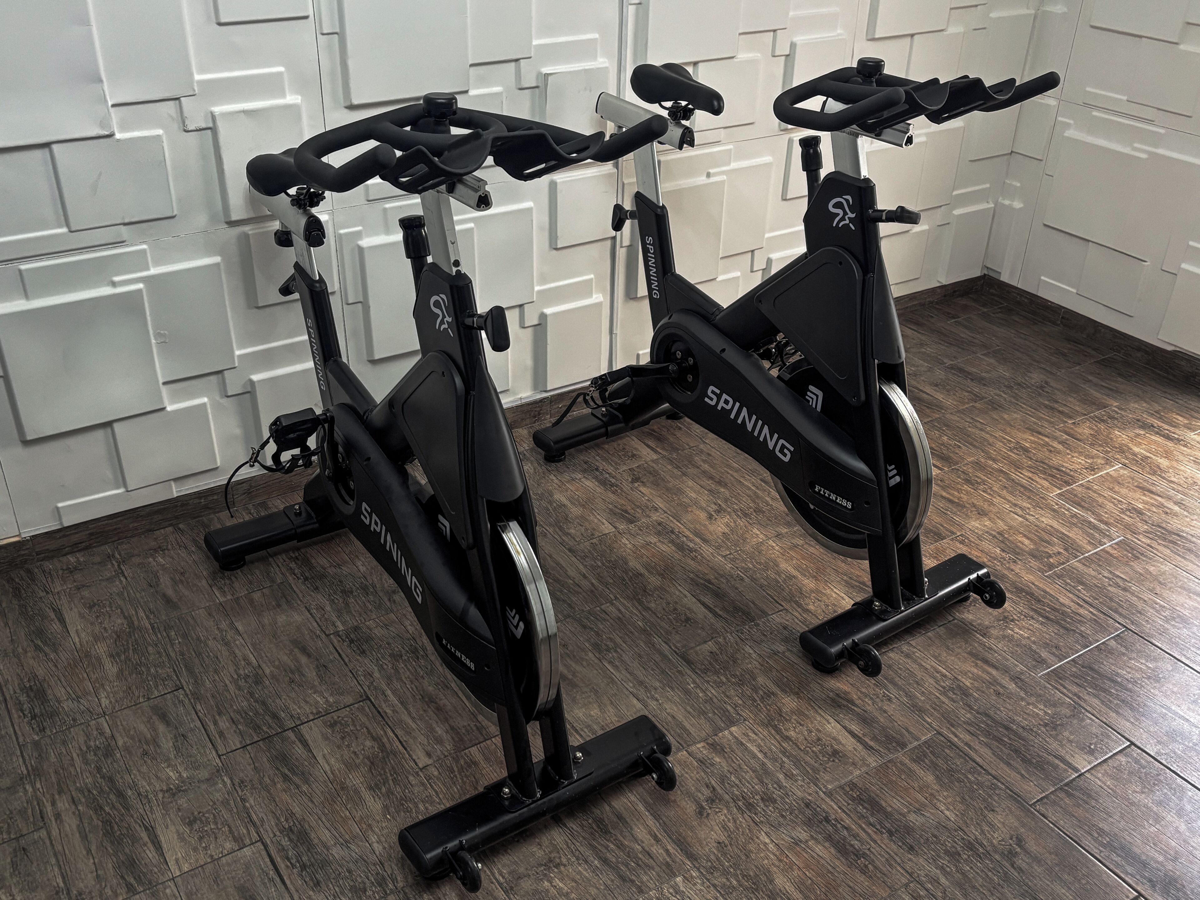 Duas bicicletas ergométricas em um quarto com parede estampada e piso de madeira.