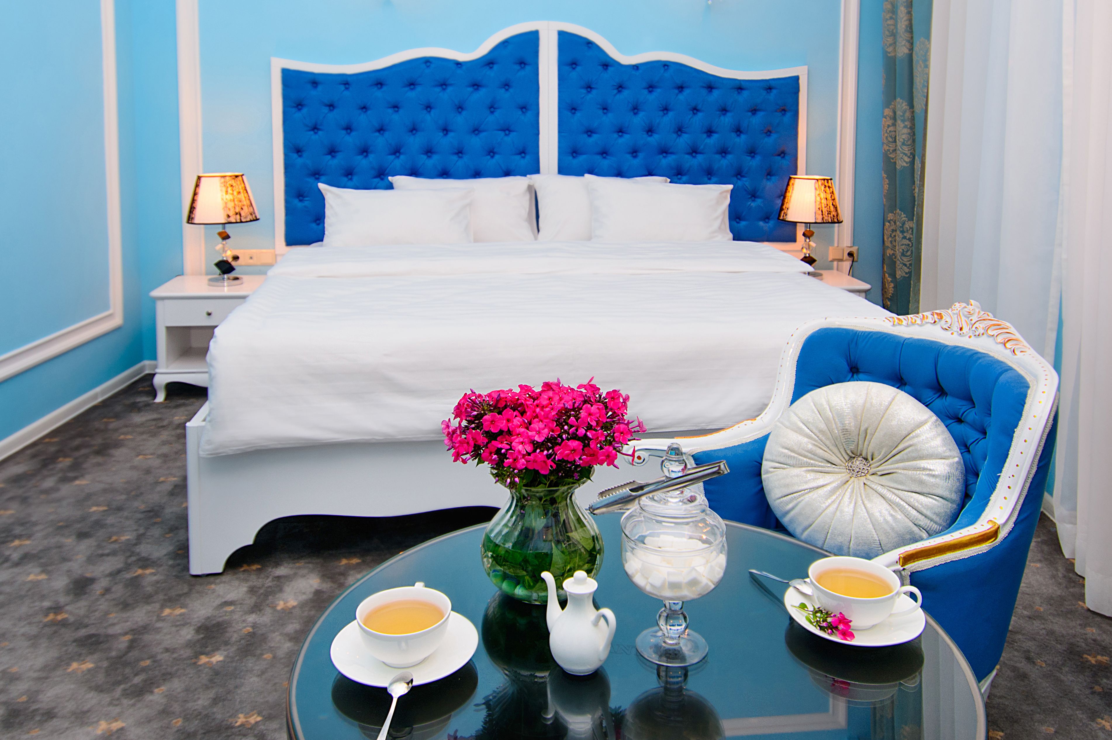 A hotel room with a large bed, a blue upholstered chair, a glass table with a vase of flowers, and two cups of tea.