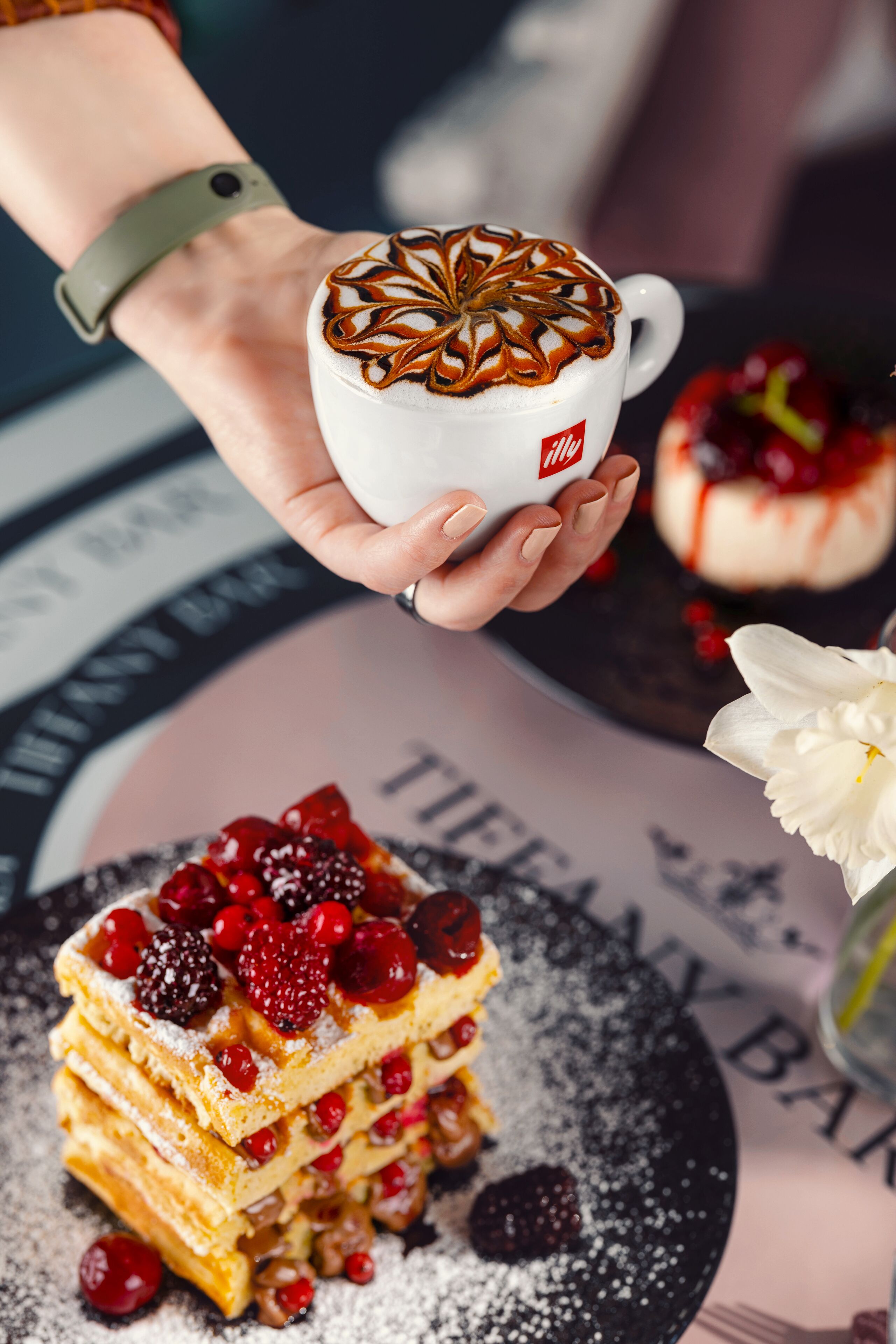 A hand holding a cup of coffee with latte art, a stack of waffles with berries, and a plate with powdered sugar.