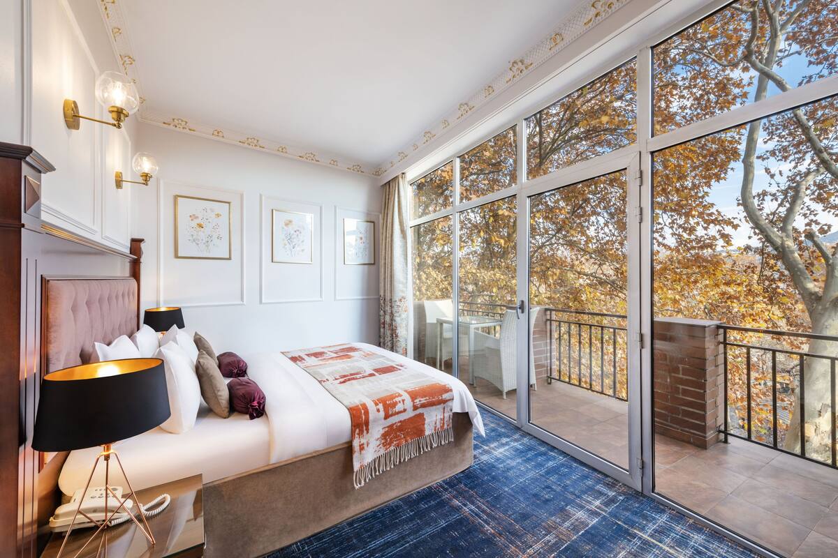 A bedroom with a large window, a bed, a nightstand, and a lamp.