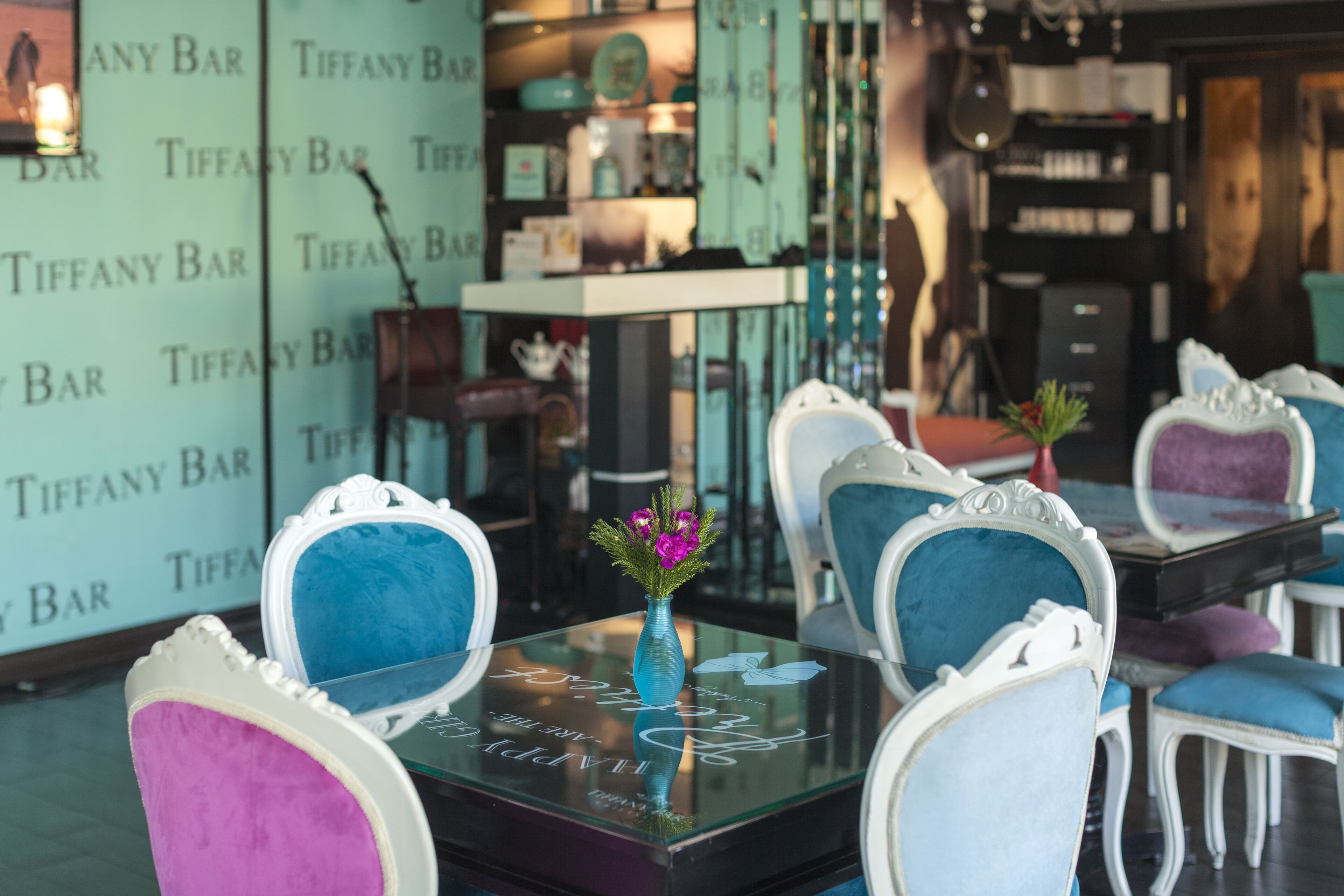 L’intérieur d’un bar Tiffany, avec des fauteuils bleu et blanc richement décorés, une table en verre surmontée d’un vase, et un décor mural portant le nom du bar.