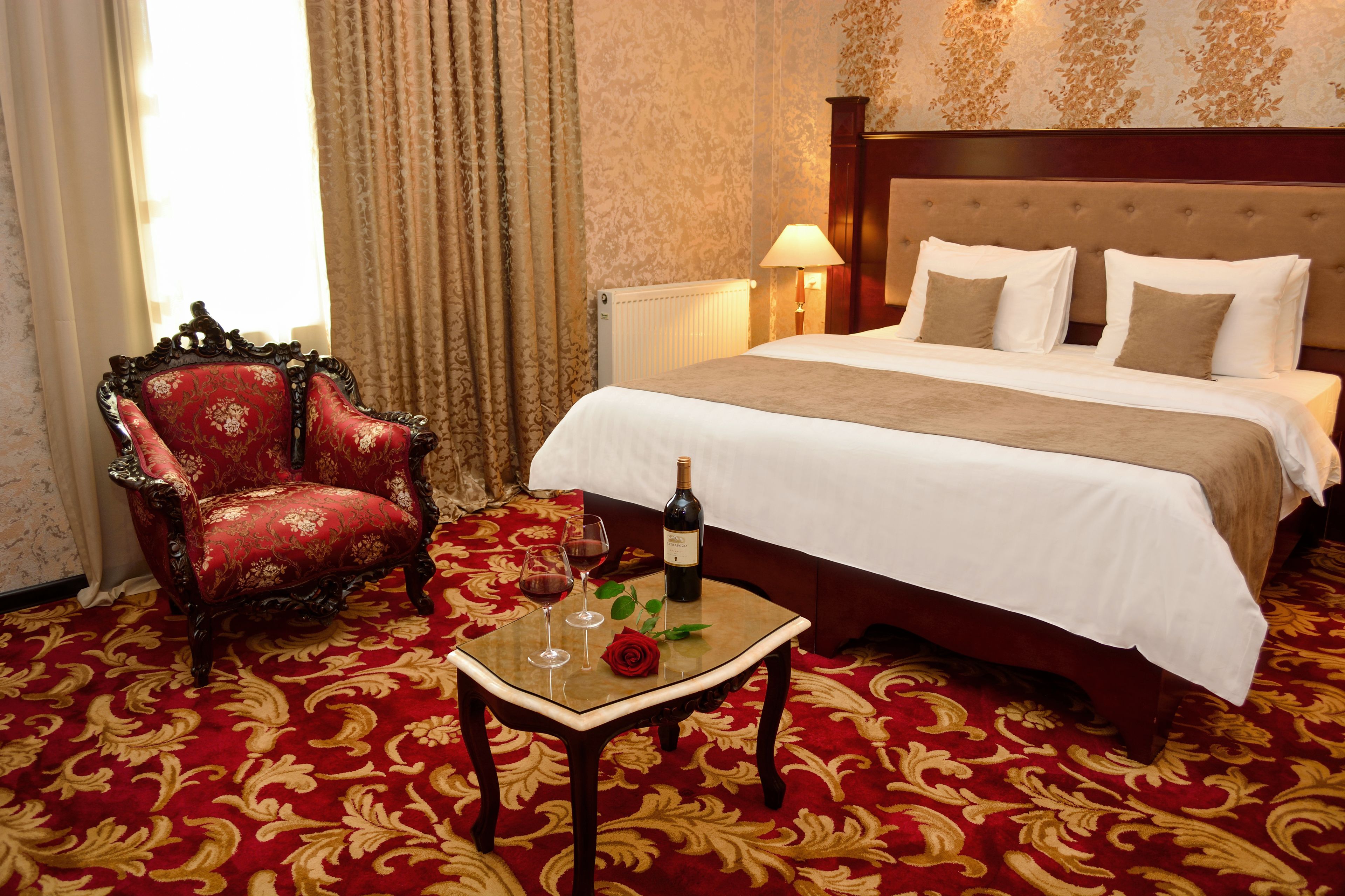 Ein Hotelzimmer mit einem großen Bett, einem roten Sessel, einem kleinen Tisch mit einer Weinflasche und Gläsern sowie einem roten Teppich.
