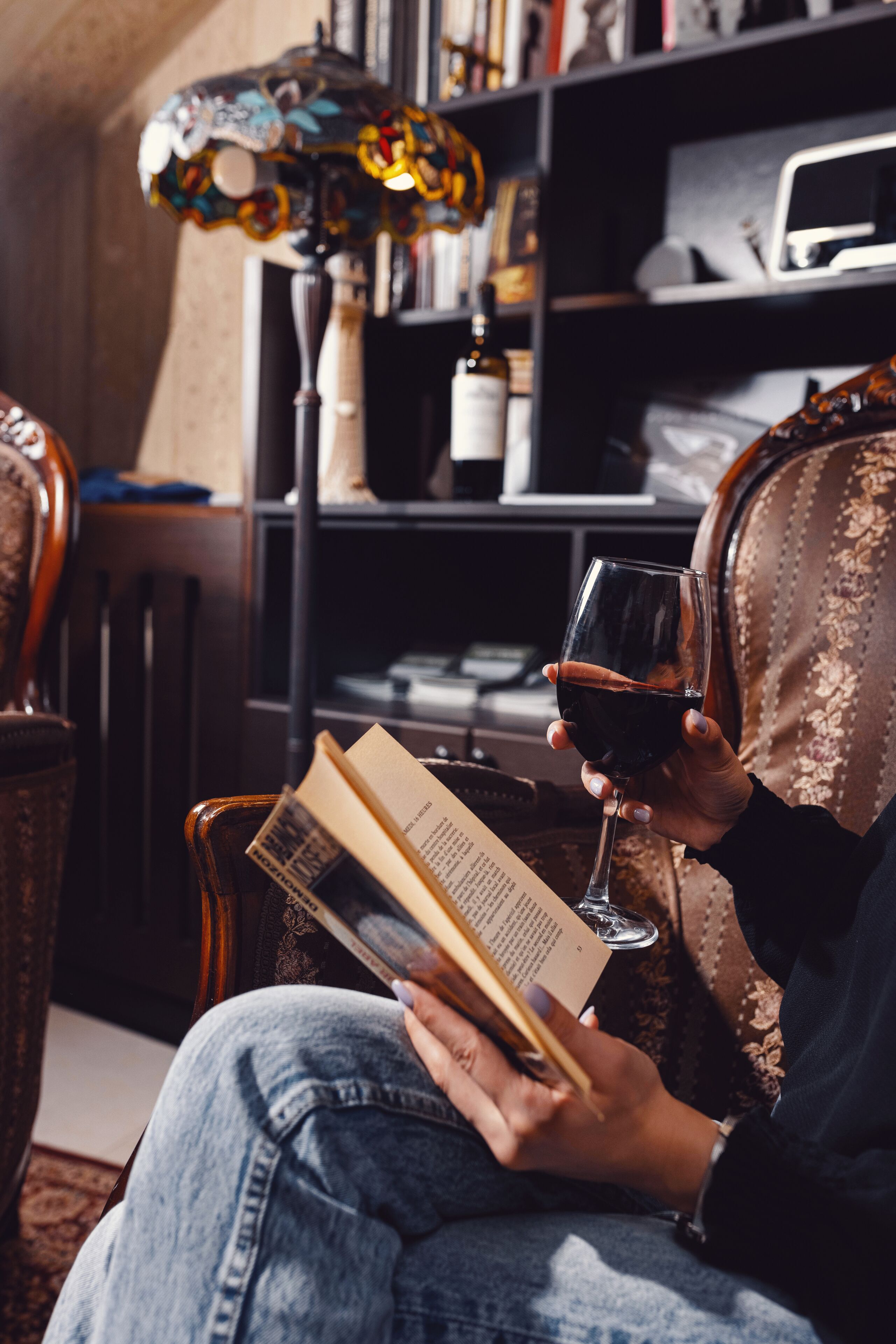 A person reading a book and holding a glass of red wine in a cozy room with a bookshelf and a lamp.