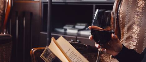 A person reading a book and holding a glass of red wine in a cozy room with a bookshelf and a lamp.