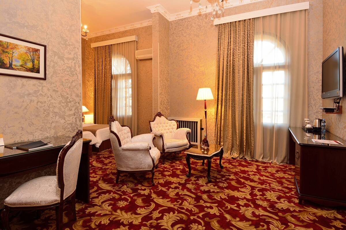 A hotel room with a red carpet, a desk with a lamp, two armchairs, a television, and a painting on the wall.