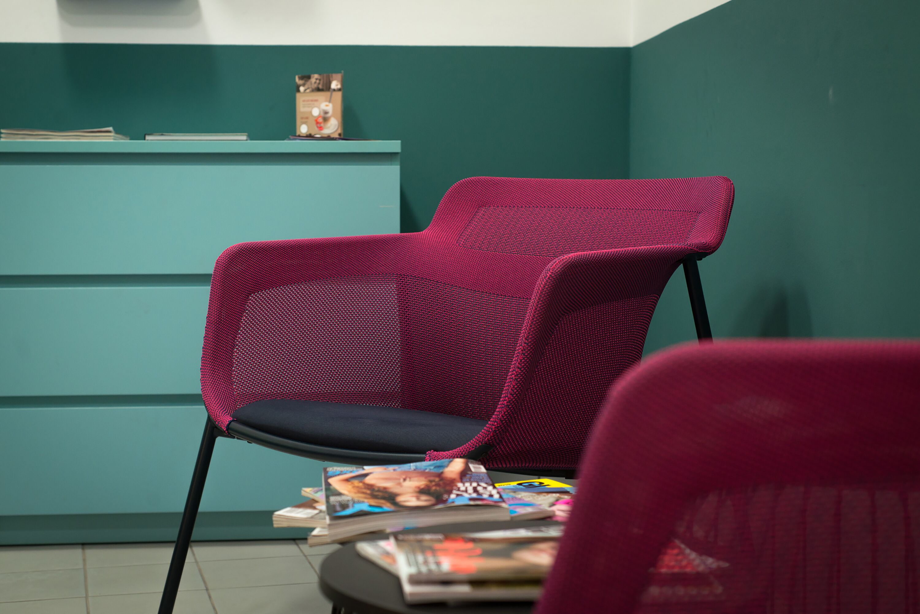 Un bureau de couleur bleu-vert, agrémenté d’une petite horloge et de quelques livres. Une chaise rouge avec un dossier en tissu maillé est placée devant le bureau.