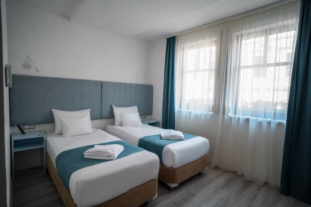 Une chambre d’hôtel avec deux lits, une table de chevet, une lampe fixée au mur et une fenêtre avec des rideaux.