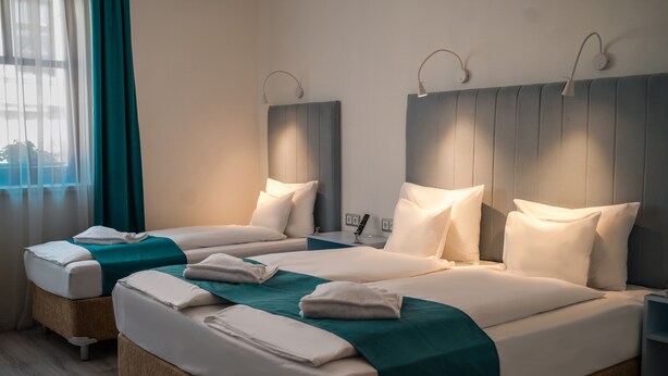 Une chambre d’hôtel avec deux lits, chacun agrémenté de draps blancs et d’une couvre-lit turquoise. Chaque lit est surmonté d’une lampe fixée au mur, et la chambre dispose d’une grande fenêtre avec des rideaux.