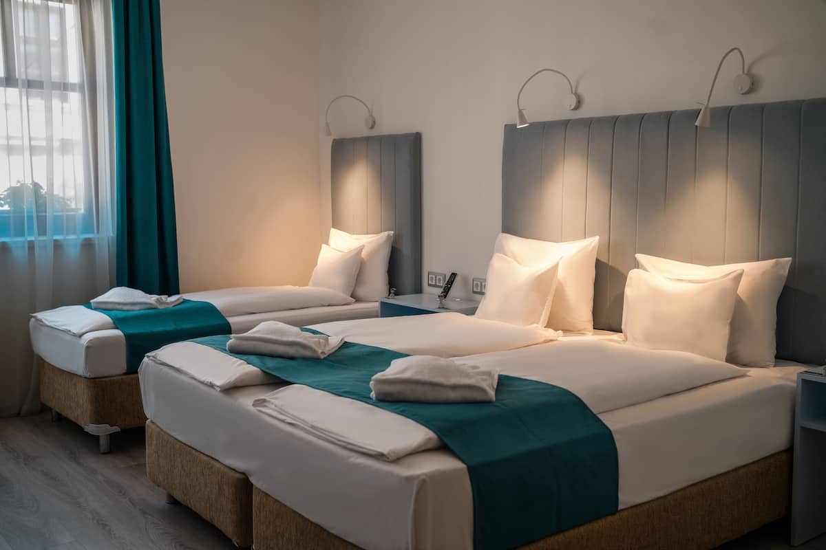 Une chambre d’hôtel avec deux lits, chacun agrémenté de draps blancs et d’une couvre-lit turquoise. Chaque lit est surmonté d’une lampe fixée au mur, et la chambre dispose d’une grande fenêtre avec des rideaux.