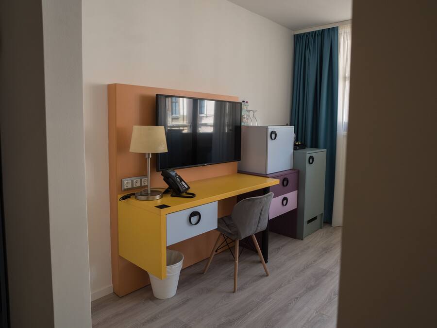 A compact hotel room with a yellow desk, a grey chair, a flat-screen TV, a small fridge, and a bedside table with a lamp.