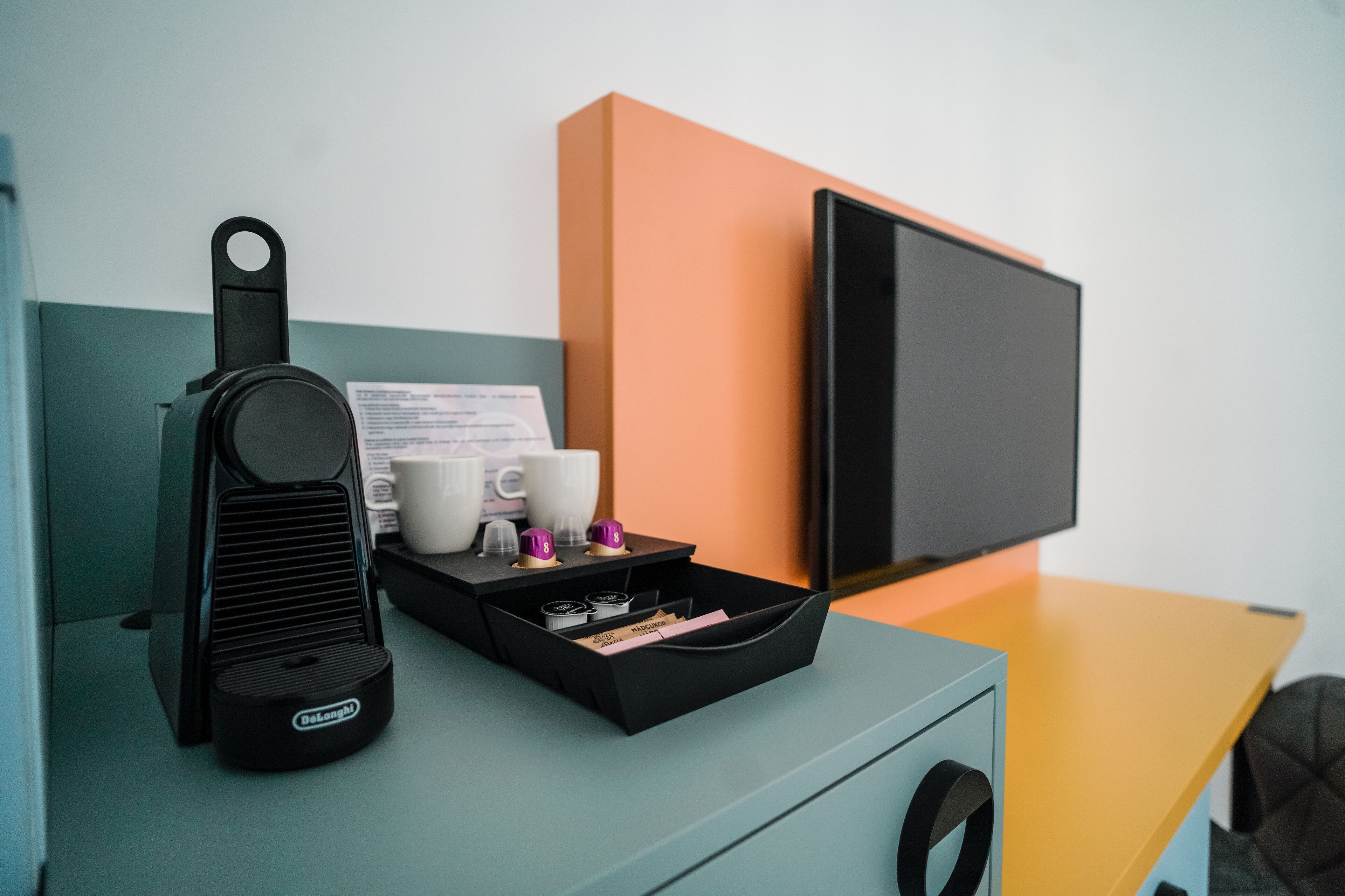 A modern room with a teal desk, a black speaker, a tray with coffee cups and a menu, and a TV on an orange wall.