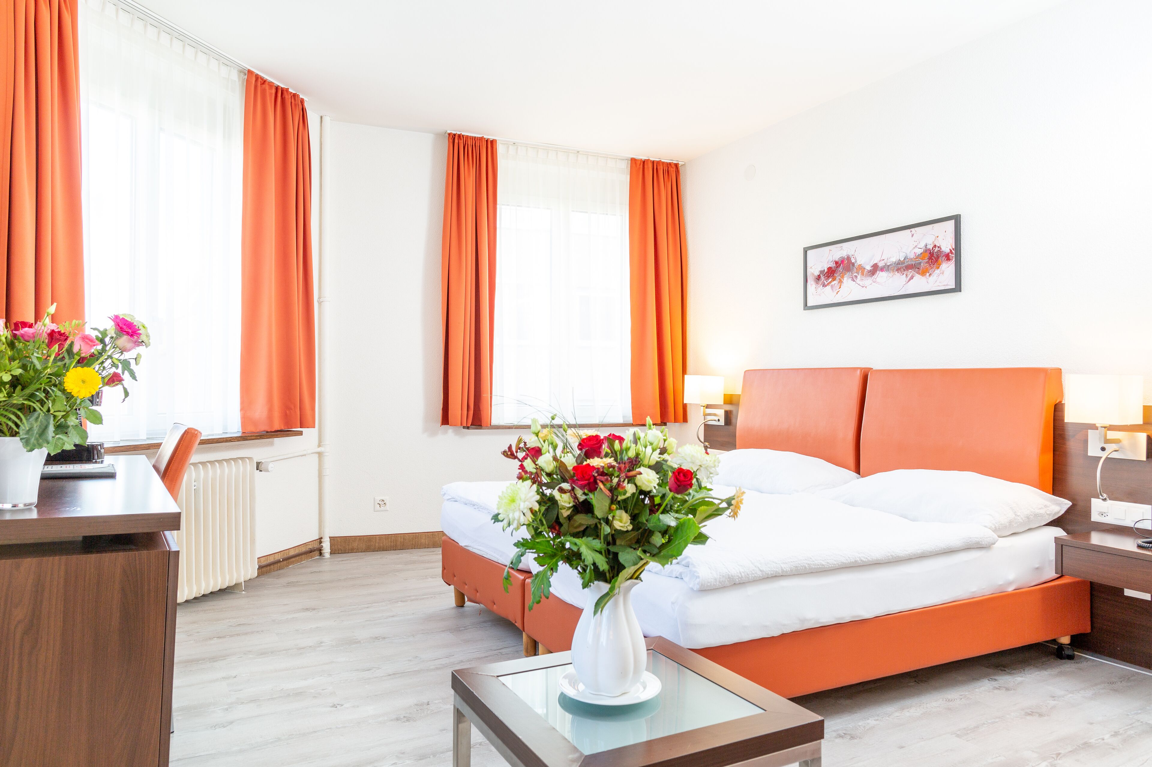 Une chambre d’hôtel avec un lit, un bureau, une chaise, un vase de fleurs et une fenêtre avec des rideaux.