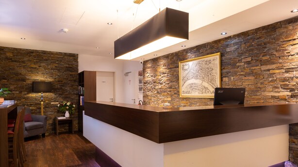 A reception area with a stone wall, a wooden reception desk, a sofa, and a lamp. - Hotel Rheinfelderhof (Basel)