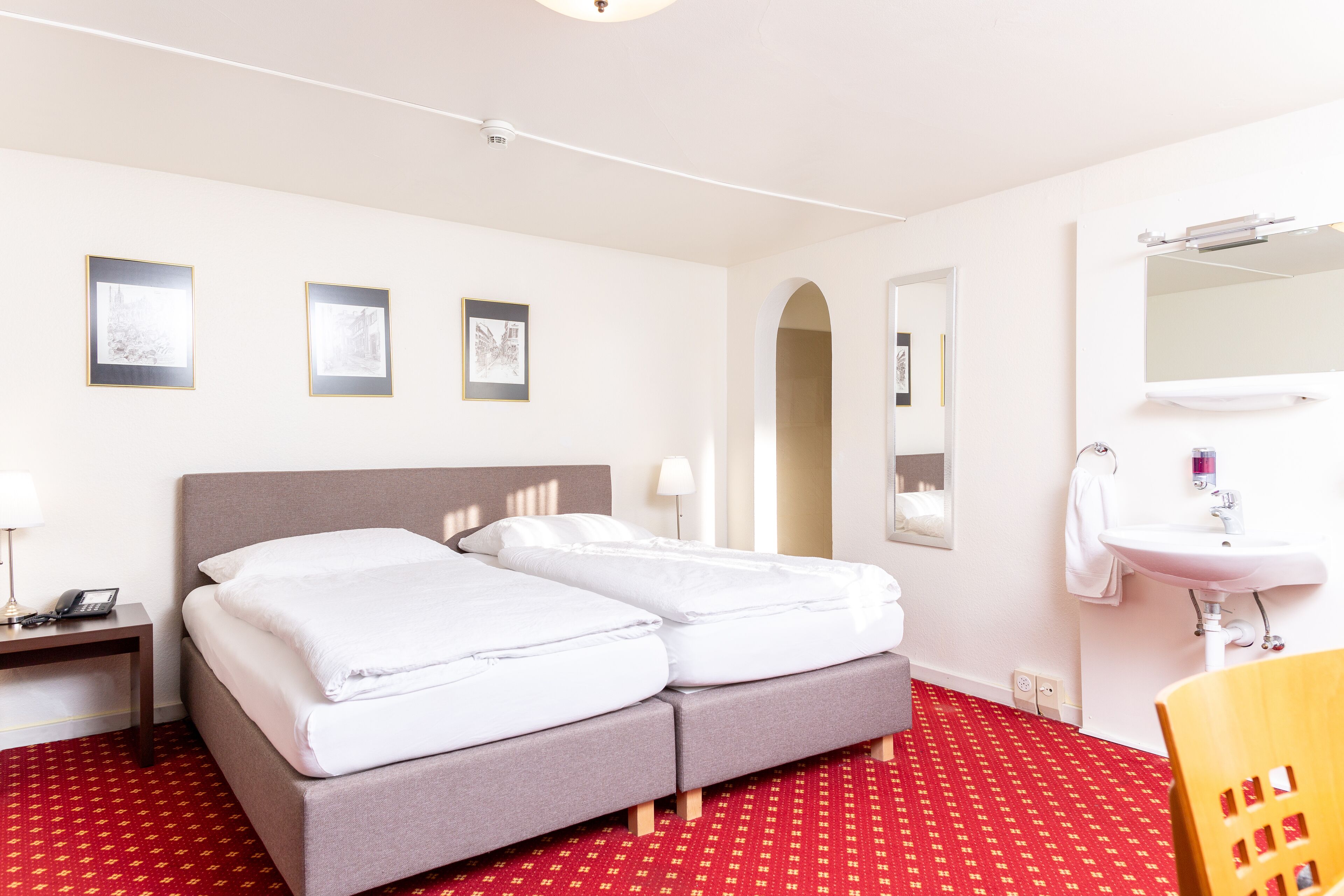 A hotel room with a double bed, a bedside table with a lamp, a red patterned carpet, a wooden chair, and a bathroom with a sink and mirror.