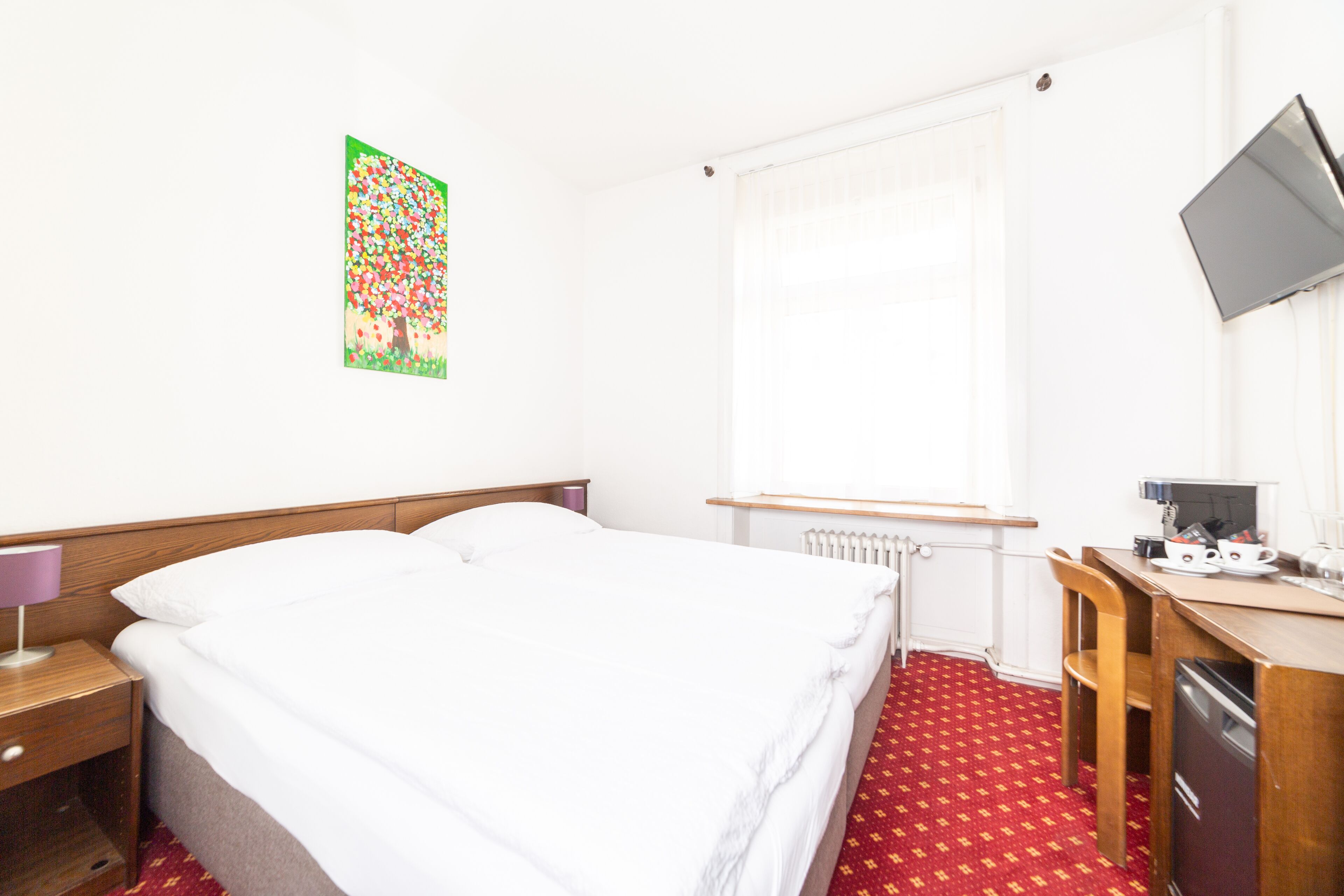 Habitación de hotel con cama, escritorio con ordenador, radiador y ventana con cortinas.