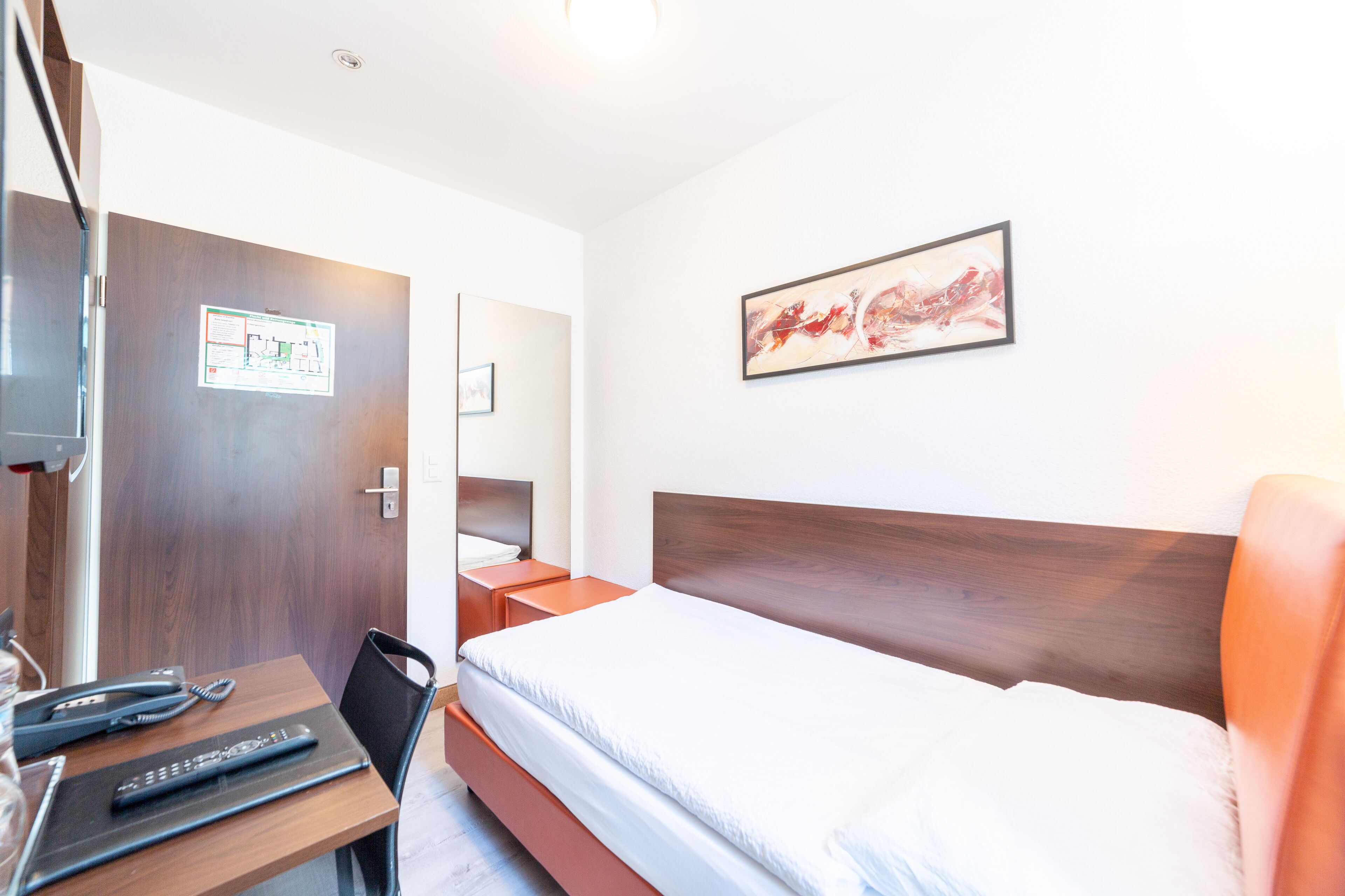 A hotel room with a bed, a desk with a telephone and remote control, a mirror, and a framed abstract painting on the wall.