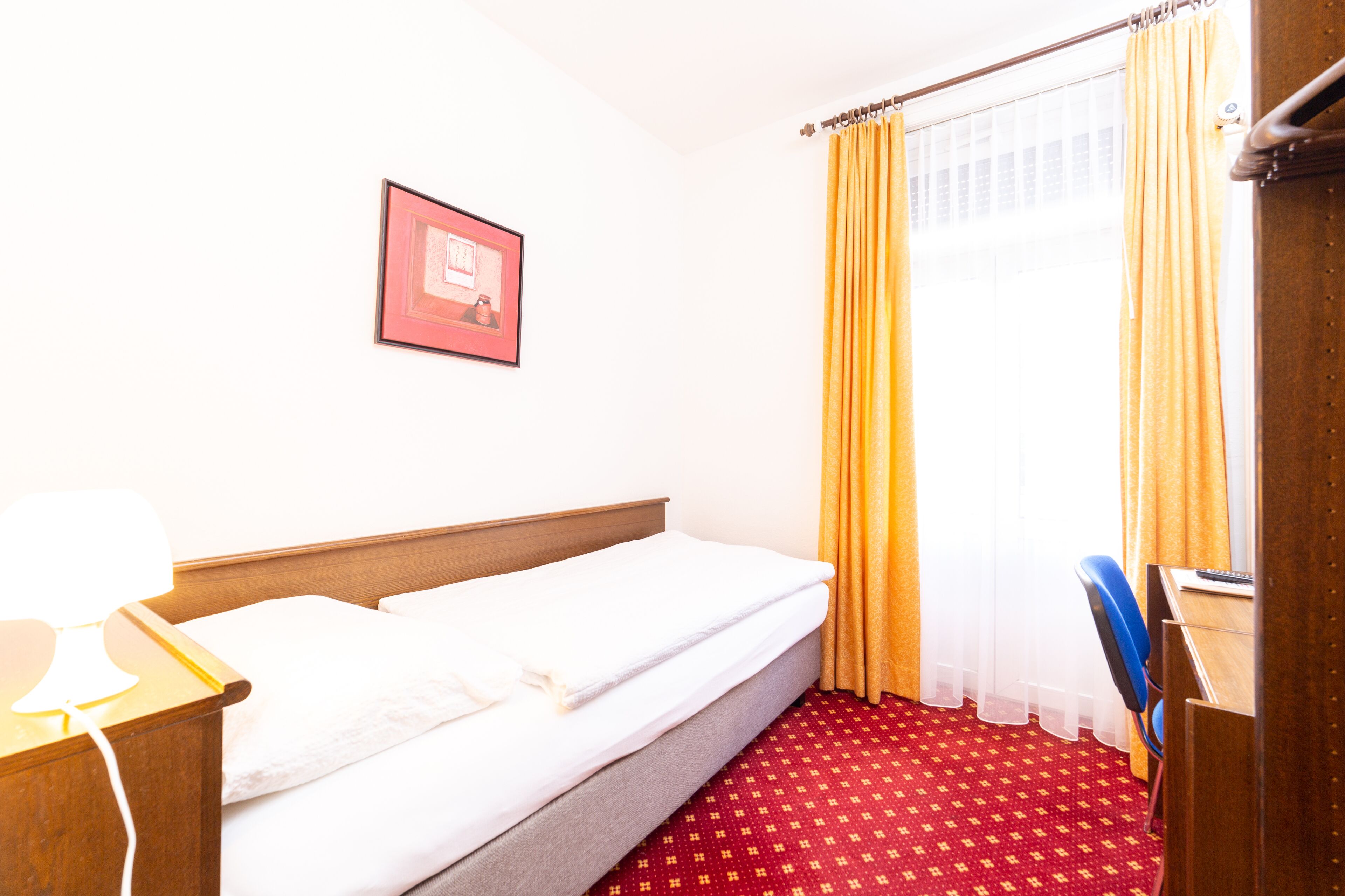 Habitación de hotel con cama, escritorio con silla, un cuadro en la pared y una ventana con cortinas.