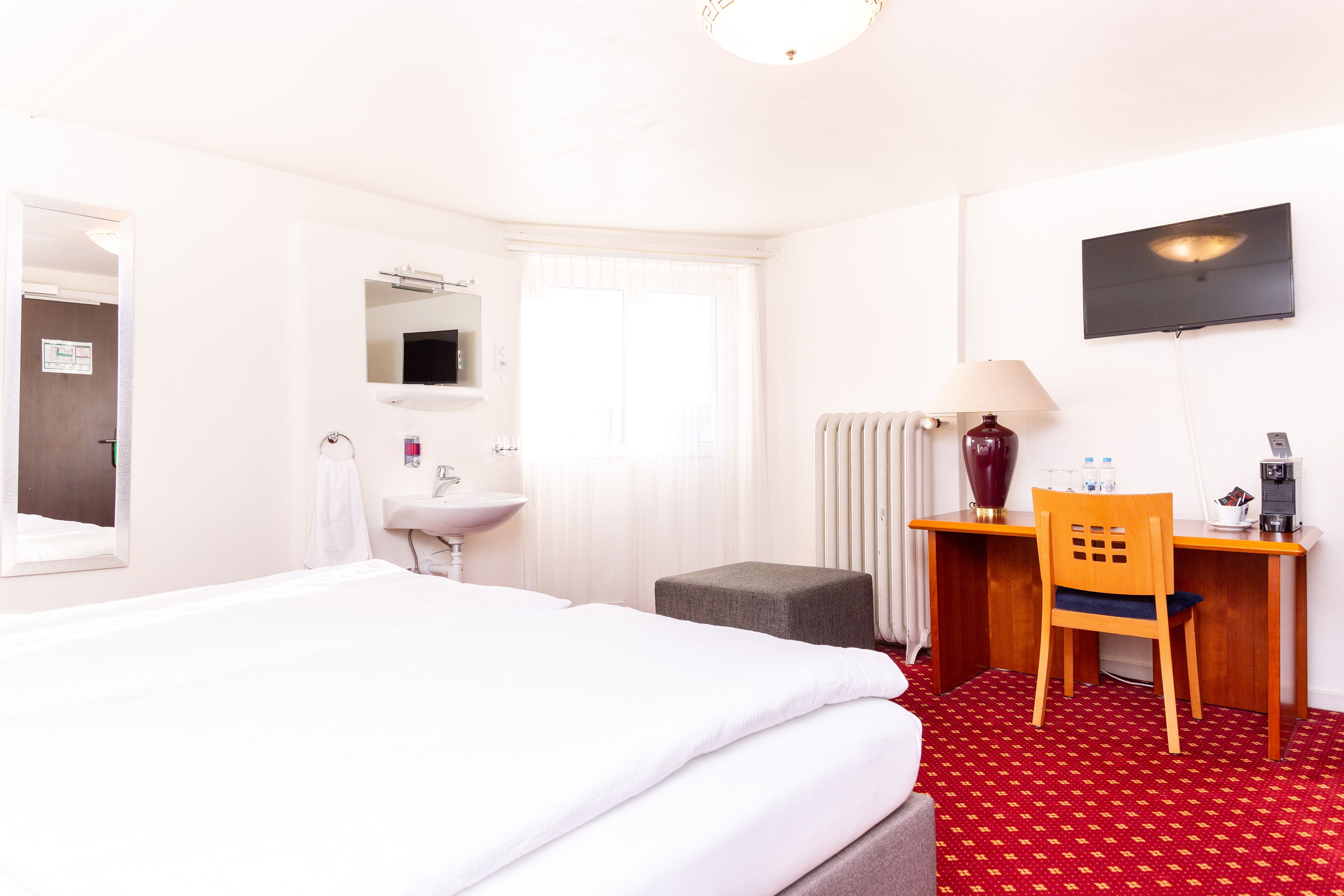 A hotel room with a bed, a desk with a chair, a TV, a lamp, and a radiator.